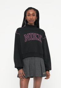 Melnā saīsinātā džemperis ar rozā Nike logo, ribinātām piedurknēm un augstu apkakli; savienots ar pelēku plisētu svārku ar smalkām šuvju detaļām.