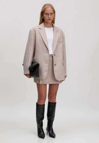 Hellbeige Blazer, passender Rock und weißes Oberteil. Schwarze strukturierte Tasche und knielange schwarze Stiefel mit spitzem Fuß. Glatte Texturen.