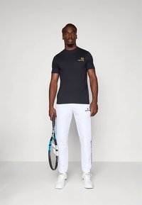 Melna īsroku sporta krekls ar dzeltenu logotipu, kopā ar baltām sviedru biksēm ar sānu akcentiem, turot tenisa raketi. Baltas sporta kurpes.