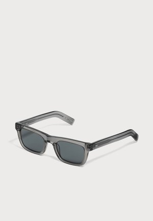 Lunettes de soleil rectangulaires gris foncé avec des montures épaisses translucides et des verres foncés sur fond blanc.