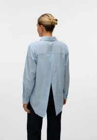 Svetlo modra srajca z dolgimi rokavi in gumbi, z odprtino na hrbtni strani in koničastim ovratnikom. Mehka tkanina in sproščen kroj, v kombinaciji s temnimi kavbojkami.