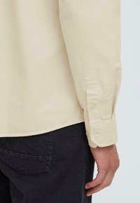 Camisa de botones de color crema con cuello puntiagudo y mangas largas, que presenta puños abotonados. Se combina con pantalones oscuros, mostrando la textura de la tela.