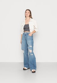 Casaco de ganga branco, top cropped preto com padrão, jeans claros de perna larga, desgastados, e sandálias brancas com solas pretas.