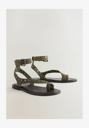Olivově zelené sandály s perforovanými semišovými popruhy a stříbrnými hroty, s otevřenou špičkou a plochou černou podrážkou. Zapínání na sponu kolem kotníku.