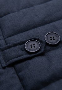 Dva černá tlačítka označená "OMBRE" na tmavě modré polstrované tkanině s viditelným prošíváním a rybí kostí vzorem.