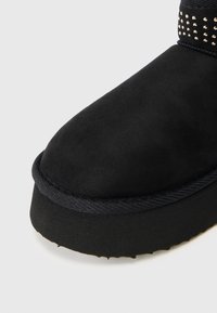 Bota negra até ao tornozelo com uma textura de camurça suave, bico arredondado, sola preta em contraste e ornamentada com detalhes metálicos na parte superior.