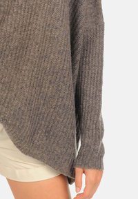 Pull en tricot gris avec texture côtelée, coupe ample et manches longues, présentant un dos plus long et un détail d'ouverture sur le côté.
