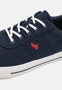 Navy canvas sneaker with white laces, round eyelets, and a red embroidered bird logo. Features a rubber sole with a contrasting stripe.