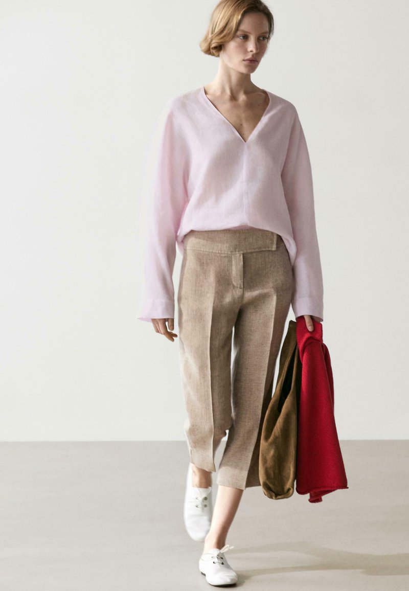 Jonge vrouw die loopt, gekleed in een lichtroze V-hals trui, beige cropped broek, witte veterlaarzen, terwijl ze rode en bruine jassen vasthoudt.