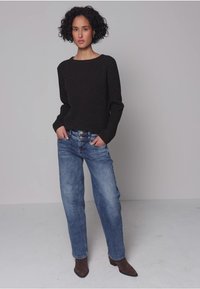 Zwarte textuursweater, lichtblauwe jeans met wijde pijpen en bruine enkellaarsjes. Model staat met handen in de zakken tegen een neutrale achtergrond.