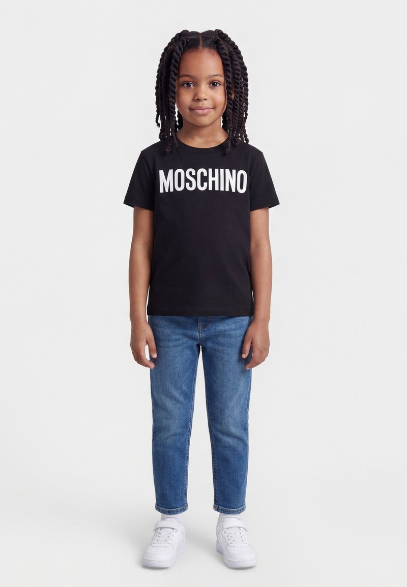 Νεαρή κοπέλα με πλεξούδες, που φοράει μαύρο t-shirt Moschino, μπλε τζιν και λευκά αθλητικά παπούτσια, στέκεται μπροστά σε ένα απλό φόντο.