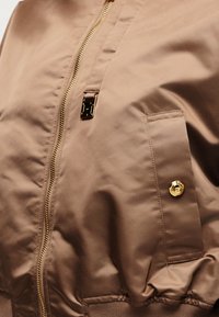 Κοντινό πλάνο ανοιχτό καφέ μπουφάν με χρυστό φερμουάρ, τσέπη με καπάκι και χρυσό κουμπί, και μεταλλική λεπτομέρεια λογότυπου κοντά στο φερμουάρ.