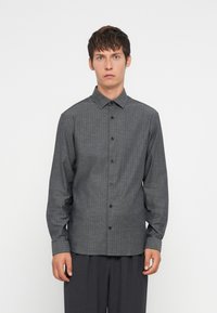 Camisa de manga larga gris con patrón, cuello con botones, botones negros y un corte relajado, combinada con pantalones oscuros.