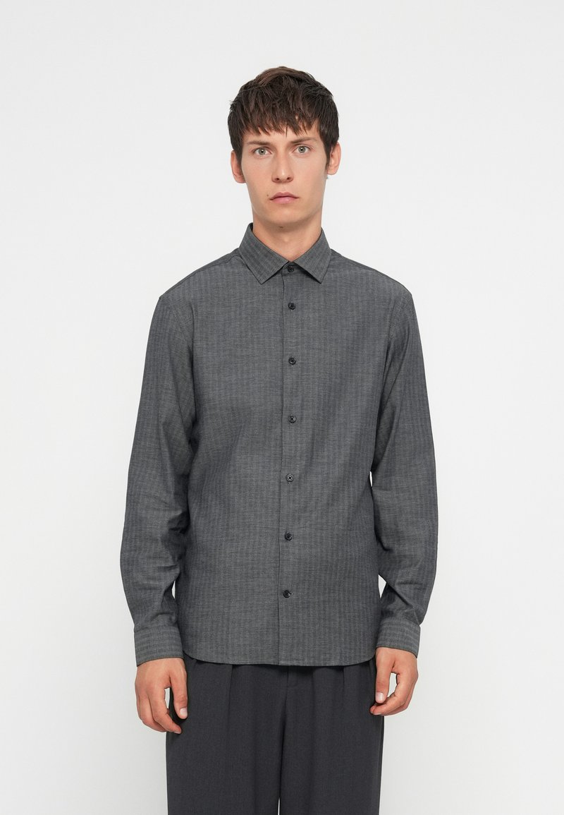Camisa de manga larga gris con patrón, cuello con botones, botones negros y un corte relajado, combinada con pantalones oscuros.