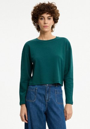 Mujer con cabello corto y rizado, vistiendo una camiseta de manga larga verde oscuro y vaqueros azules de cintura alta, de pie frente a un fondo liso.