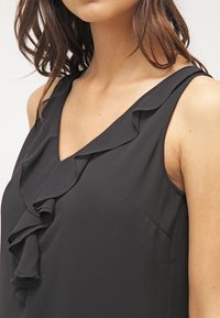 Kobieta w czarnej bluzce bez rękawów z dekoltem w serek oraz falbaną wzdłuż dekoltu i przodu.