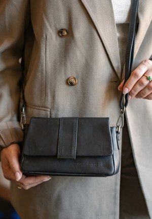Person trägt einen beigen Blazer und hält eine schwarze Leder-Umhängetasche mit genähten Details und Klappenverschluss.