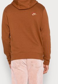 Sudadera de algodón marrón con capucha con cordón y logo de Nike bordado en la parte posterior. Combinada con pantalones de pana claros a rayas.