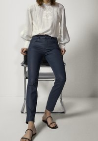 Marinafärgade byxor med slät yta, hög midja och femficksdesign, parat med en vit blus med dekorativ broderi och vida ärmar.