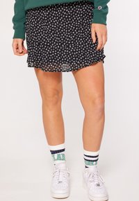 Person som bär en svart blommönstrad mini-kjol, grön sweatshirt, vita strumpor med ränder och text, samt vita sneakers, som står mot en enkel bakgrund.