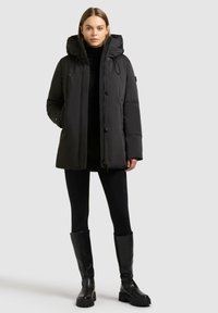 Zwarte pufferjack met capuchon, voorzien van een rits aan de voorkant en knopen, zijzakken en een gladde textuur. Gedragen over zwarte leggings en laarzen.
