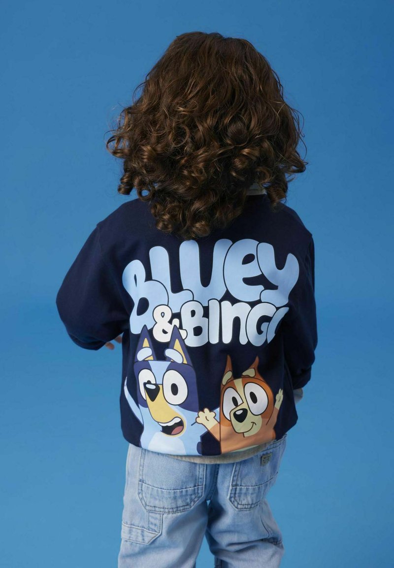 Marineblå genser med store trykk av tegneseriehundene Bluey og Bingo på baksiden, med hvit tekst over. Laget av bomull, med rund hals.