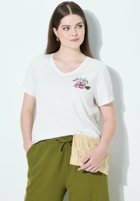 Camiseta blanca de cuello en V con bordado de flores rosas y hojas verdes, combinada con pantalones verde oliva y un bolso de paja trenzada.