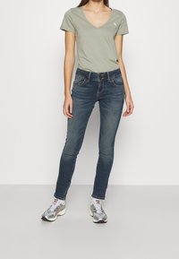 Grünes V-Ausschnitt-T-Shirt kombiniert mit dunkelblauen Skinny Jeans. Die Jeans haben verwaschene Akzente und ein klassisches Fünf-Taschen-Design. Silberne Sneakers runden das Outfit ab.