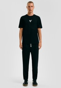 Zwarte katoenen T-shirt met korte mouwen en een klein, lichtgrijs logo. Gecombineerd met zwarte broek met elastische tailleband en zwarte sneakers.