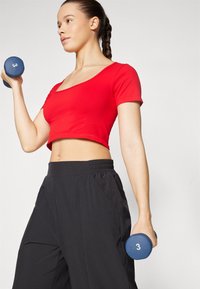 Rød kortermet crop-top kombinert med svarte, løse treningsbukser. Holder en blå hantel merket "3." Enkel design og atletisk passform.