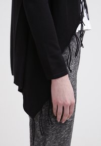 Profil latéral d'une personne portant un haut noir à manches longues avec des détails à franges et un pantalon gris texturé.