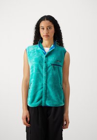Teal fleece vest med en teksturert overflate, blå detaljer, trykknapplukking og en glidelås lomme på brystet. Ermeløs design, avslappet passform.
