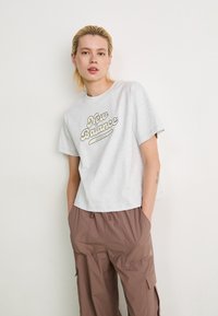 Jeune personne avec des cheveux blonds courts portant un t-shirt gris clair de New Balance et un pantalon cargo marron, se tenant devant un mur blanc uni.