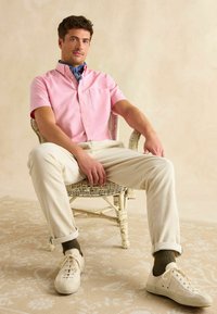 Ružová polozapínacia košeľa s vreckom, krémové kordové nohavice, biele tenisky a zelené ponožky, posedenie na tkanom kresle na vzorovanom koberci.