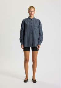 Blau gestreifte Baumwollhemd mit Knopfleiste, ausgestattet mit einer Brusttasche, langen Ärmeln und einer lockeren Passform. Kombiniert mit schwarzen Shorts und Schuhen.