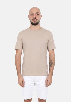 Uomo con barba e tatuaggio che indossa una maglietta beige a maniche corte e pantaloncini bianchi, in piedi davanti a uno sfondo chiaro uniforme.