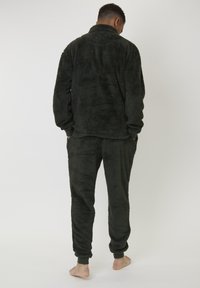 Man stående barfota med ryggen mot kameran, iförd en mörkgrön plyschfleecejacka och matchande byxor med ribbade muddar.