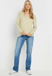 Beige Langarmoberteil mit V-Ausschnitt, kombiniert mit blauen geraden Jeans und weißen Sneakers. Der Stoff scheint weich zu sein und hat eine lockere Passform.