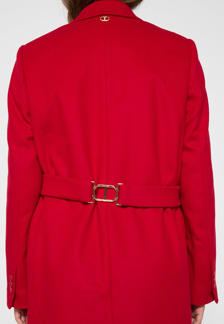 Vista posteriore di una persona che indossa un cappotto rosso con una cintura a fibbia dorata e un piccolo emblema dorato sotto il colletto.