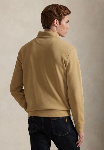 Άνδρας που φαίνεται από πίσω, φοράει μπεζ πουκάμισο με μακριά μανίκια και γιακά, και σκούρο μπλε τζιν με πορτοκαλί ραφές και μικρό κεντημένο λογότυπο.