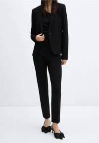 Costume noir ajusté avec une veste structurée et un pantalon slim. Accentué par de grandes chaussures à talons noirs en forme de nœud. Texture de tissu lisse.