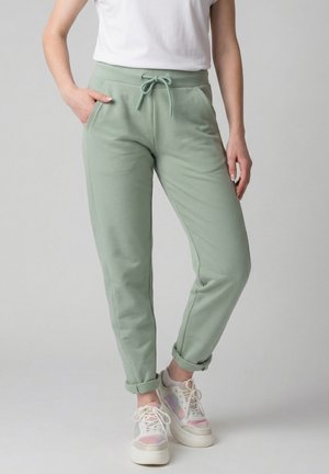 Frau trägt hellgrüne Jogginghose mit Kordelzug und hochgekrempelten Bündchen sowie weiße Sneaker, steht mit einer Hand in der Tasche.