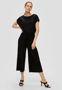 Zwarte geplooide jumpsuit met een brede halslijn, korte mouwen en een verstelbare koordsluiting in de taille. Met wijde benen en een gladde textuur.