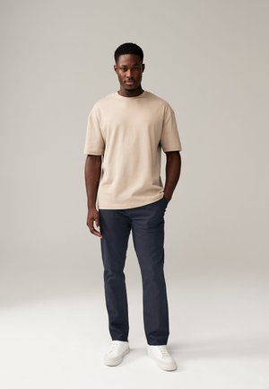 Jeune homme debout, une main dans la poche, portant un t-shirt beige, un pantalon bleu marine et des baskets blanches, sur un fond gris uni.