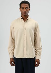 Beige overhemd met lange mouwen en een button-down kraag, gemaakt van gladde stof. Voorzien van knopen aan de voorkant en een gebogen zoom, gedragen met marineblauwe geplooide broek.