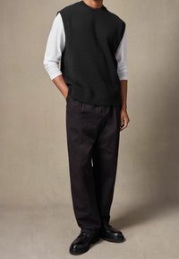 Schwarzer ärmelloser Strickpullover mit geripptem Kragen über einem weißen Langarmshirt, kombiniert mit dunklen Hosen und schwarzen Schnürschuhen.