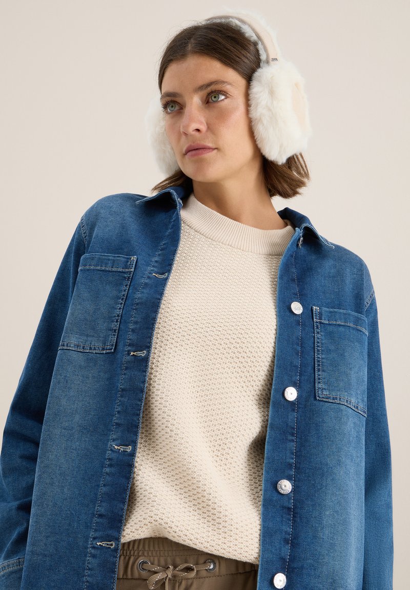 Camicia di jeans con lavaggio blu, chiusura a bottoni, taschino sul petto; maglione cremoso testurizzato sotto, abbinato a pantaloni beige e cuffie bianche soffici.