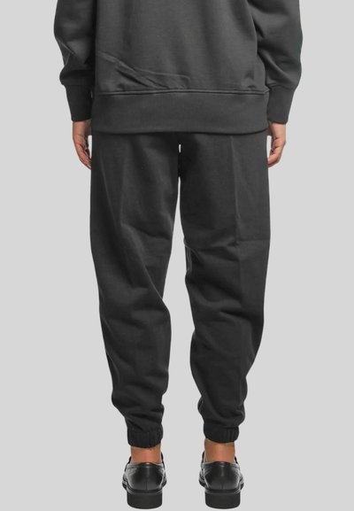 Persona che indossa pantaloni da jogging neri e scarpe nere, vista da dietro su uno sfondo grigio neutro.