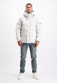 Gevoerde, lichtgrijze jas met een hoge kraag en capuchon, voorzien van een ritssluiting aan de voorkant, een borstzak en een matte afwerking. Gekleed met blauwe jeans en witte schoenen.