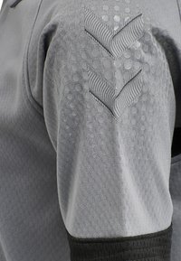 Grå idrottströja med texturerad tyg och cirkulära mönster, svarta detaljer och chevron-accenter i en mörkare nyans på ärmen.